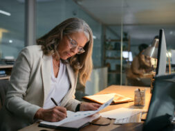 Accounting, night and writing with mature woman in office for drafting budget report at desk. Deadline, finance and notebook with professional accountant in workplace for audit, bookkeeping or tax