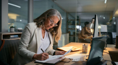 Accounting, night and writing with mature woman in office for drafting budget report at desk. Deadline, finance and notebook with professional accountant in workplace for audit, bookkeeping or tax