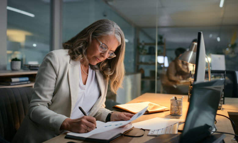 Accounting, night and writing with mature woman in office for drafting budget report at desk. Deadline, finance and notebook with professional accountant in workplace for audit, bookkeeping or tax