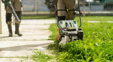 Two men are trimming grass: one is using a lawn mower, and the other one is using a blower to blow away the cut grass.
